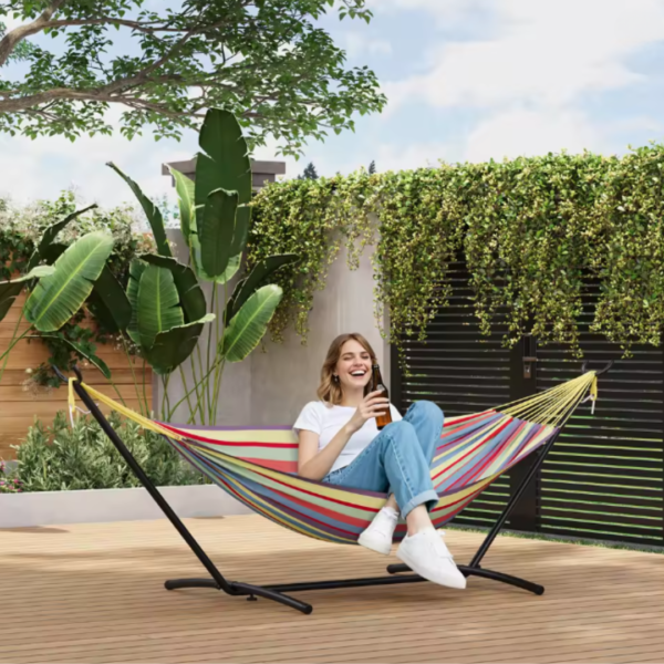 Femme souriante qui se détend avec une boisson dans un hamac avec support multicolore au cœur d'un jardin arboré.