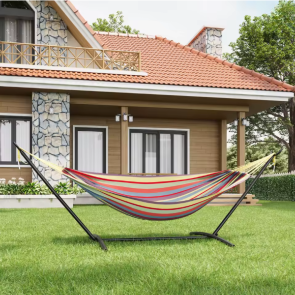 Hamac avec support en métal noir et toile à rayures multicolores installé sur une pelouse verte devant une grande maison.